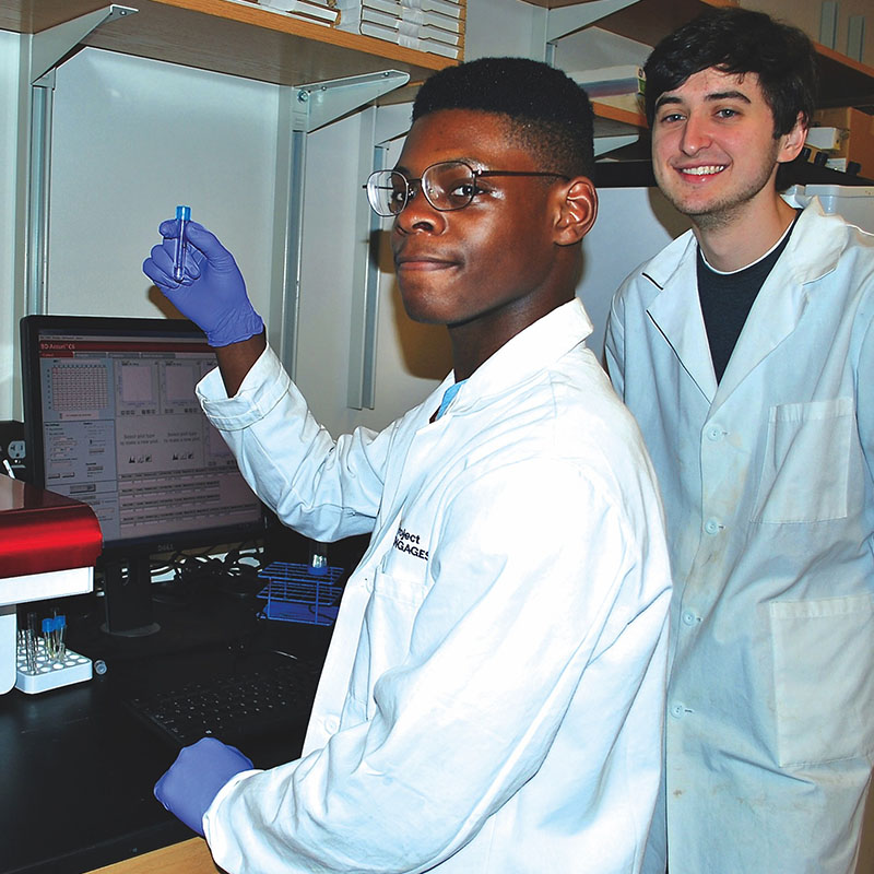 A person in a lab coat and gloves holds up a small sample vial while working at a computer displaying scientific data, with another person in a lab coat standing nearby in the laboratory.