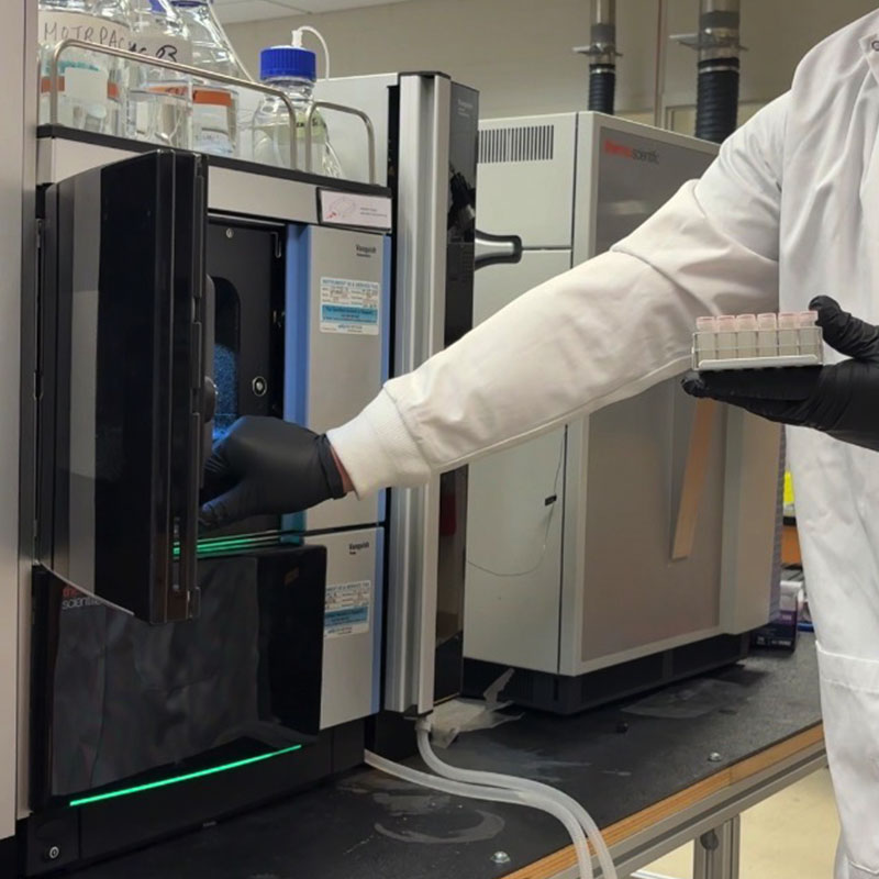A laboratory worker wearing a white lab coat and black gloves is placing a sample into an open compartment of a large analytical instrument on a lab bench. The worker holds a rack of small sample vials in one hand while reaching toward the instrument with the other.
